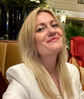 Smiling woman with long blonde hair wearing a white jacket in a warmly lit indoor setting.