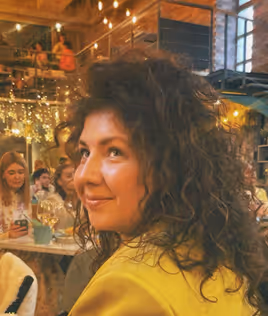 Curly-haired woman in a yellow jacket smiling in a warmly lit restaurant with patrons in the background.