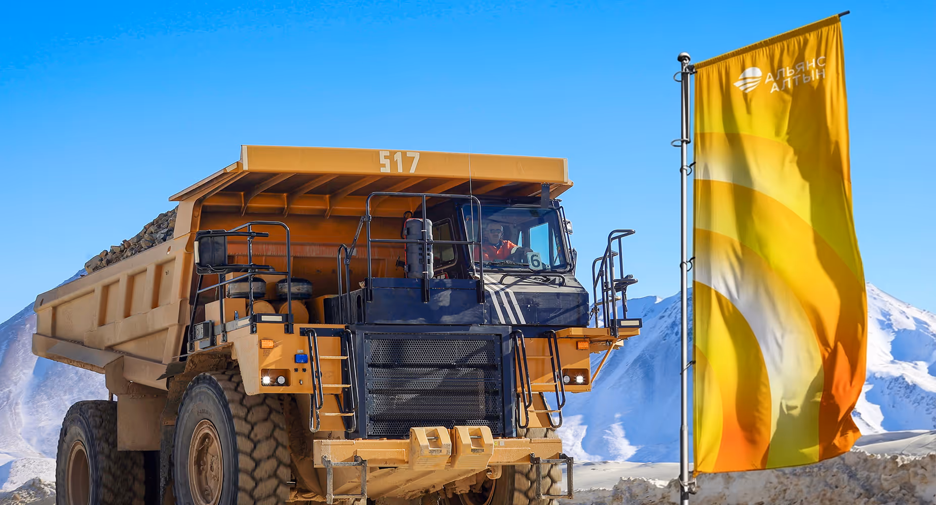 Large mining dump truck loaded with rocks near a yellow flag against snowy mountains.