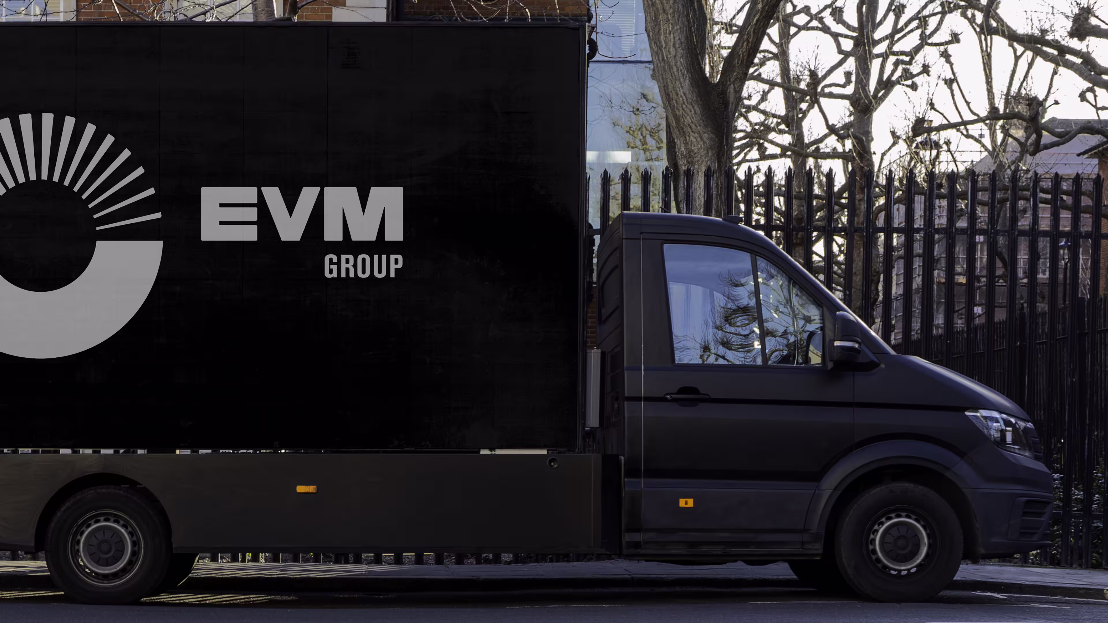 Black delivery truck with EVM Group logo parked on street with leafless trees and fence behind.