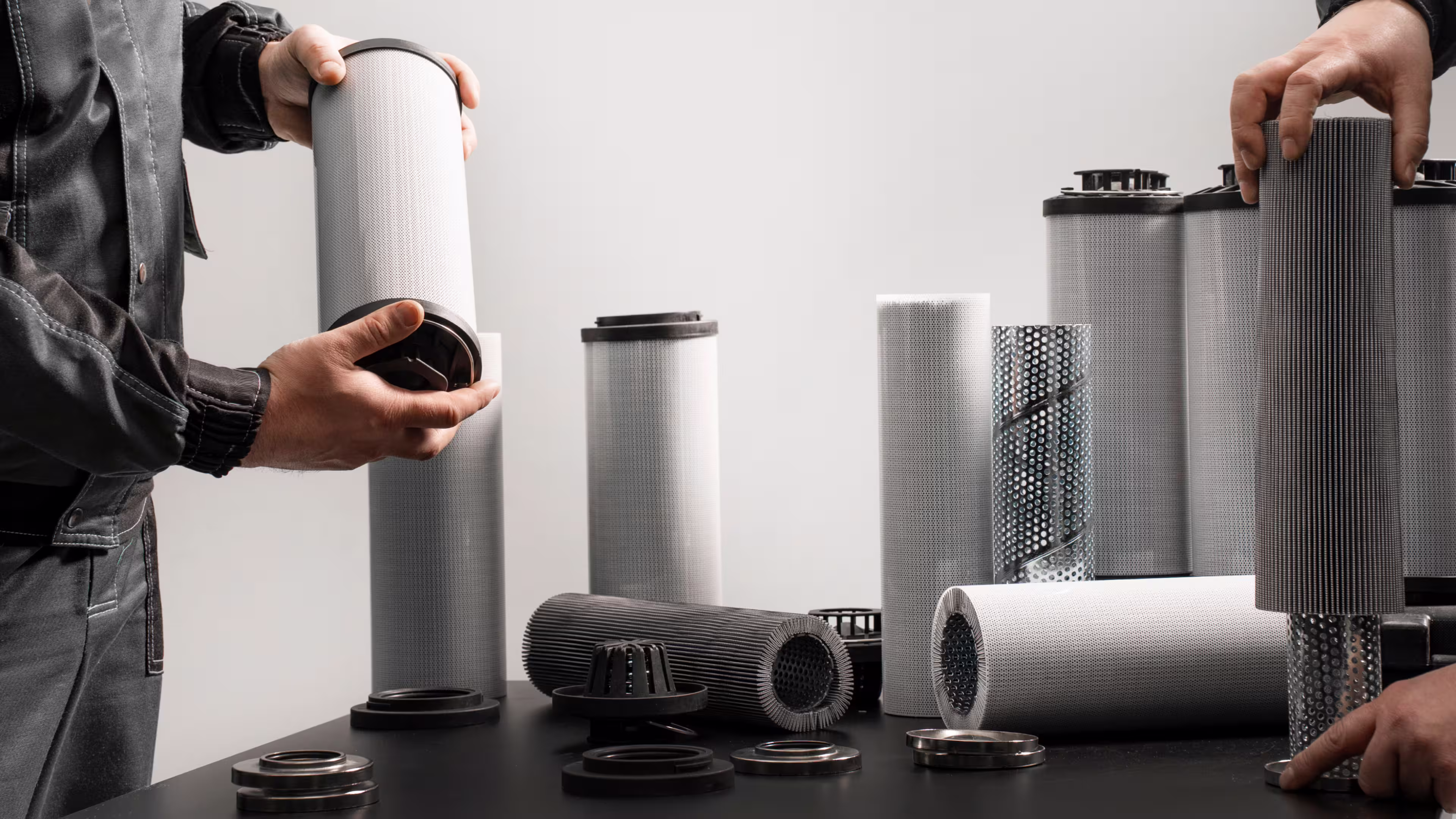 Two people handling cylindrical industrial filters and parts on a black table against a plain background.