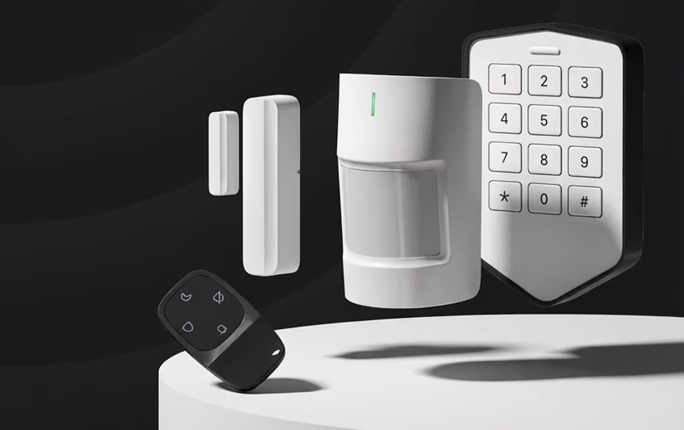 Home security system components including remote, motion sensor, keypad, and door/window sensor on a white platform.