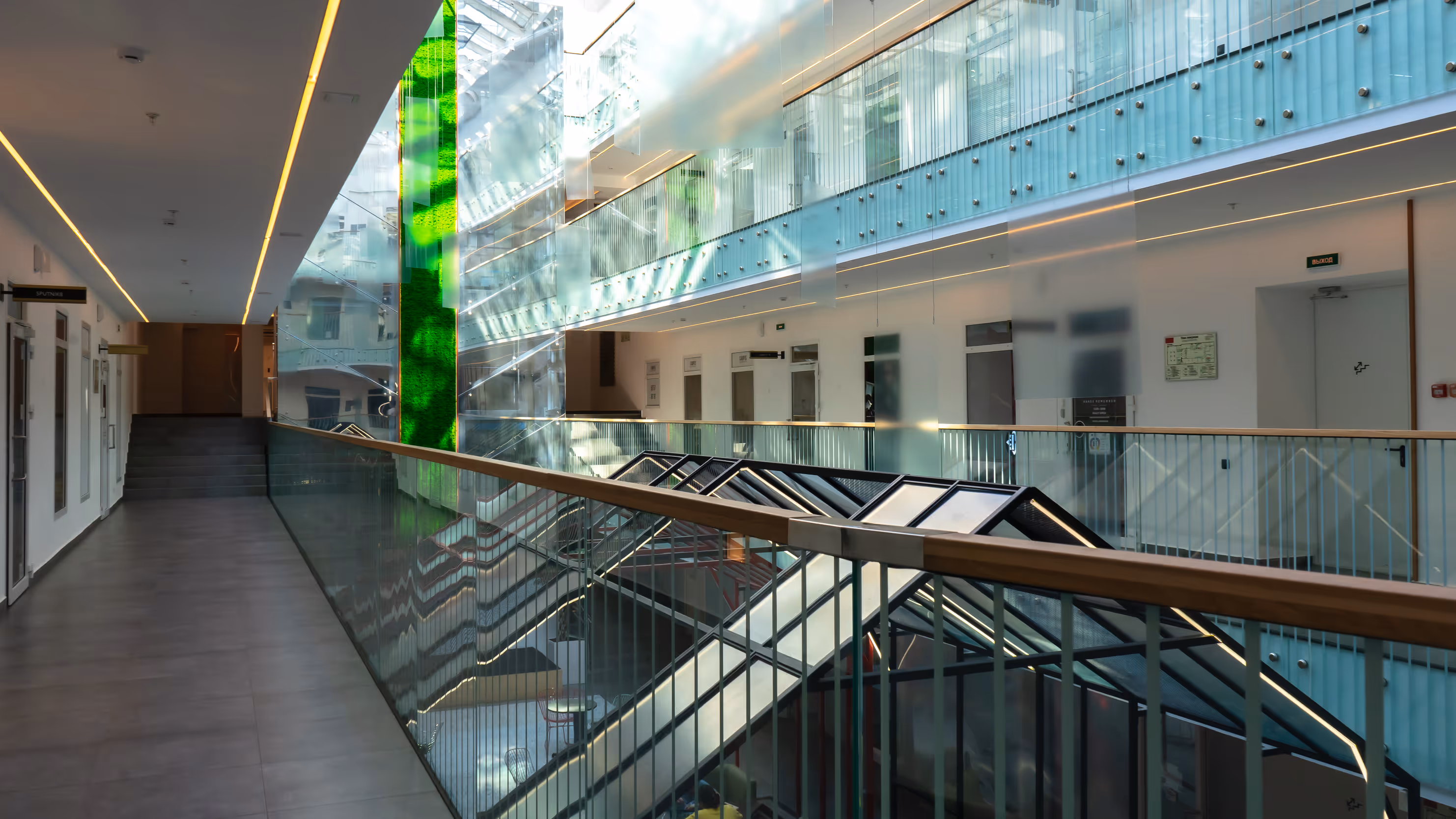 Interior corridor of modern building with glass railings, escalators, and a vertical green wall feature.