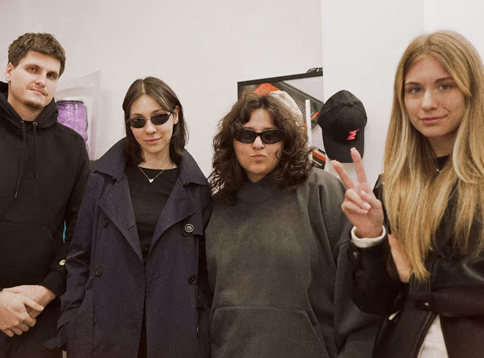 Four young adults posing indoors, two wearing sunglasses, one making a peace sign with fingers.