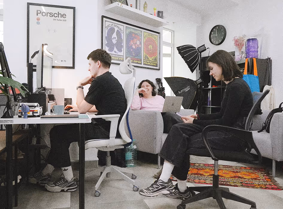 Three people working and relaxing in a creative office with computers, chairs, and wall art.