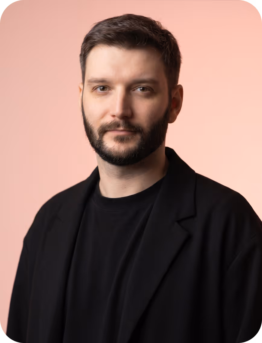 Man with short dark hair and beard wearing a black shirt and jacket, pink background.