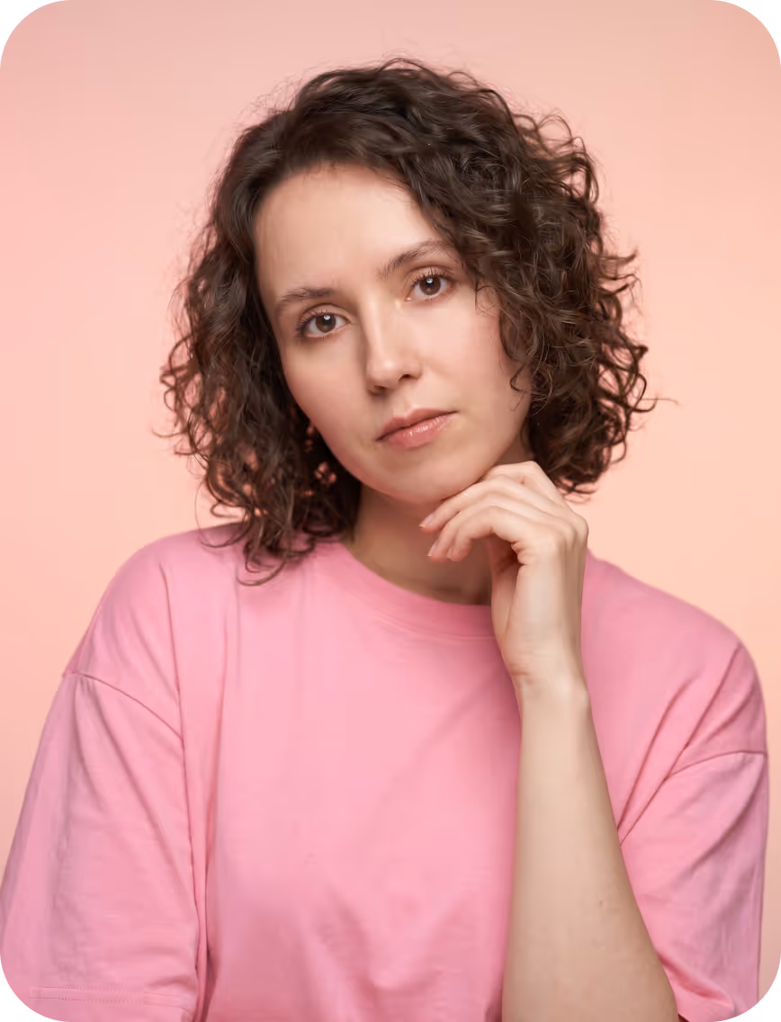 Young woman with curly hair resting chin on hand wearing pink shirt against soft pink background.