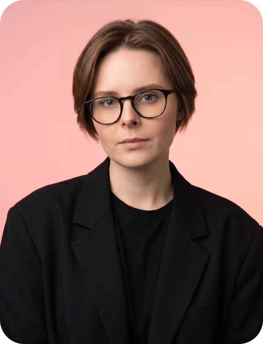 Woman with short brown hair and glasses wearing a black blazer against a pink background.
