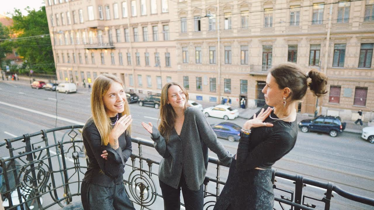 Three young women talking and smiling on a balcony overlooking a city street with buildings and cars.
