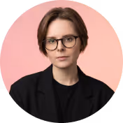 Person with short brown hair, glasses, and black blazer against a pink background.