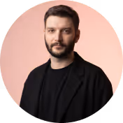 Man with short dark hair and beard wearing black shirt and jacket on light pink background.