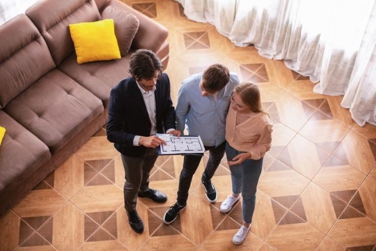 Man en vrouw staan met makelaar in huis naar plattegrond te kijken (van boven).