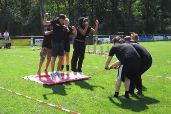 mensen trekken andere mensen op een zeil door het gras als onderdeel van een Zeskamp