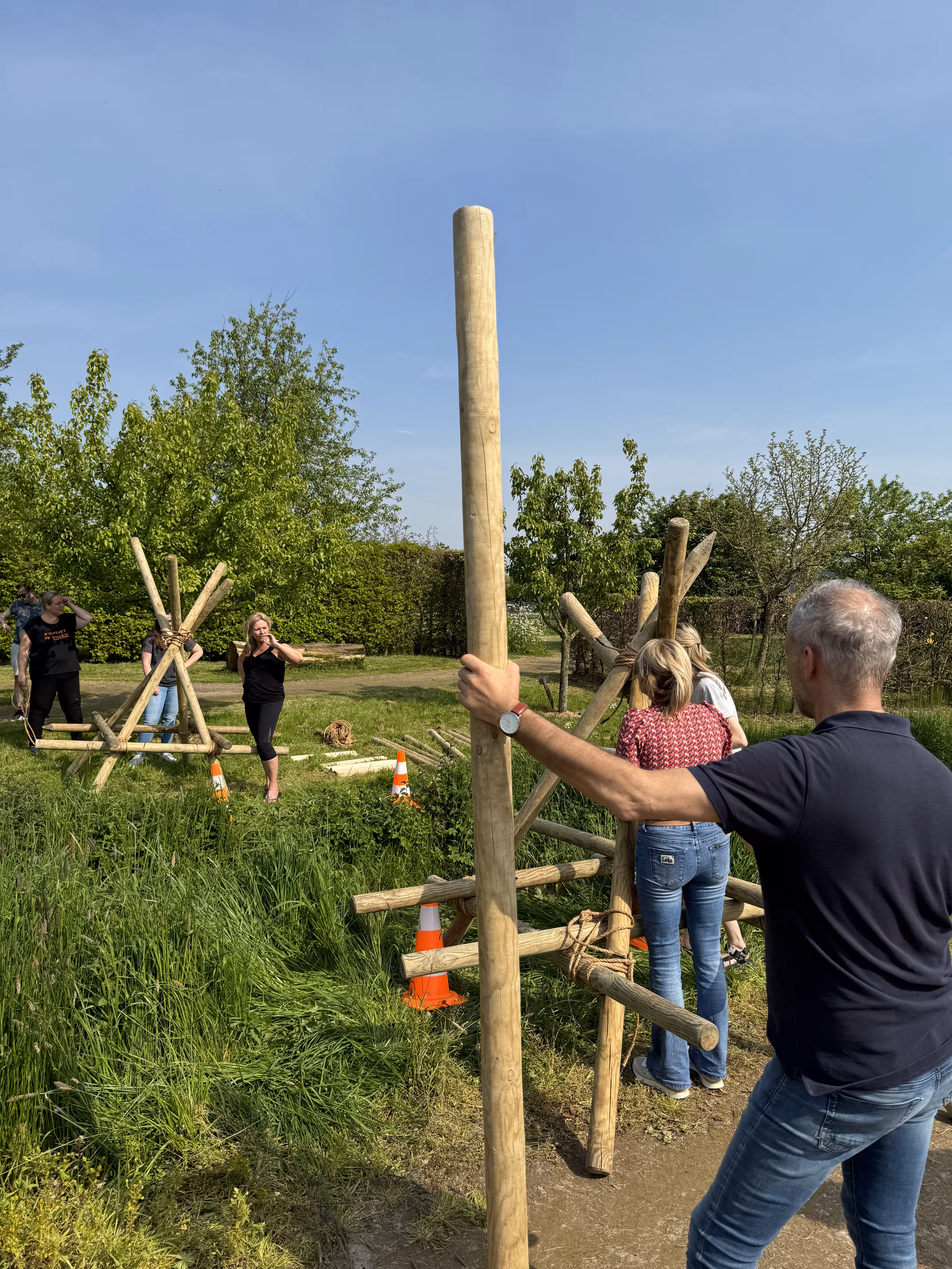 Een man houdt een houten balk vast en kijkt toe naar het team dat aan het overleggen is tijdens de workshop brug bouwen