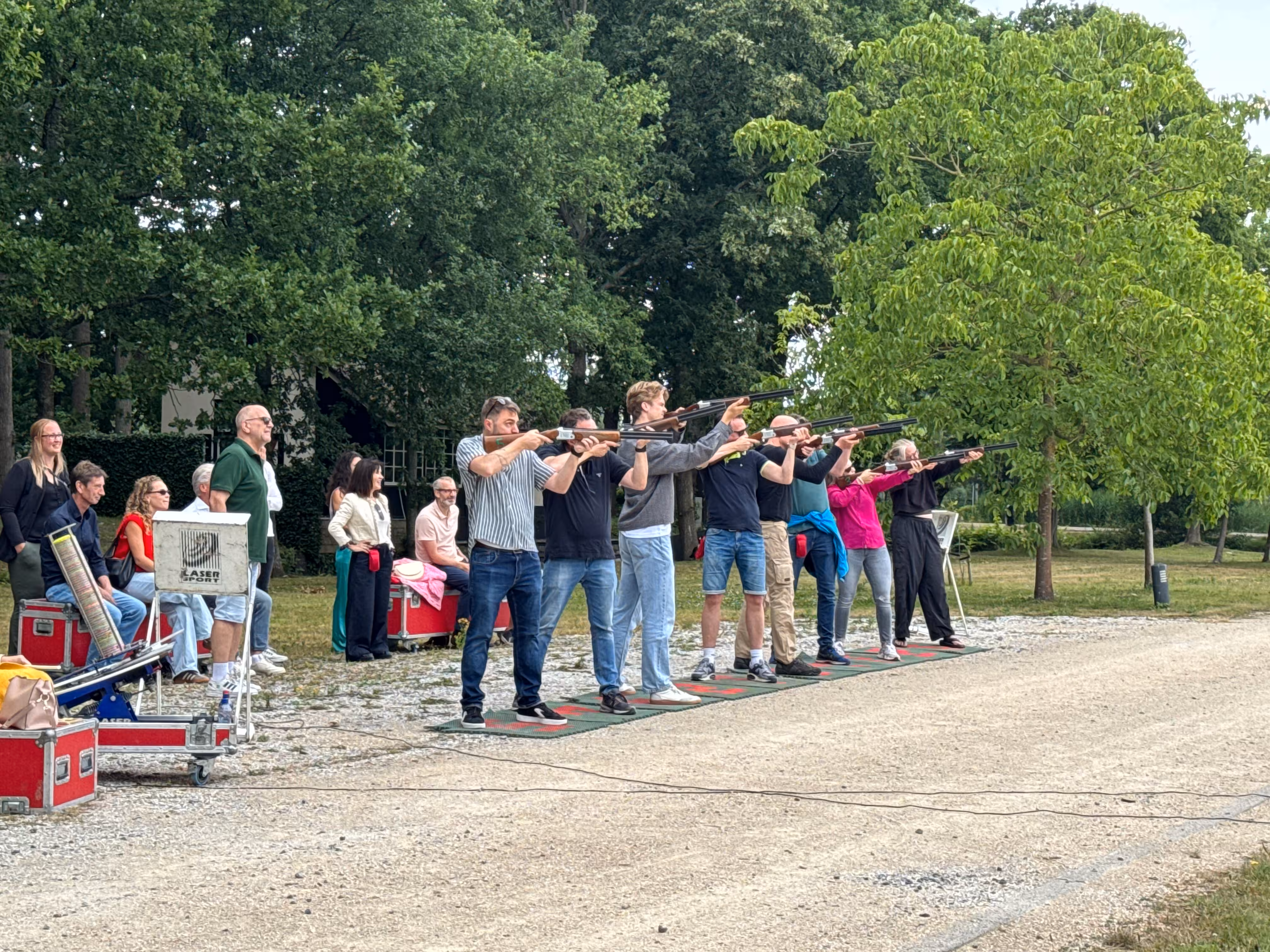 Op een grindpad staan een aantal mensen op een rij en richting met het geweer naar een laser kleiduif