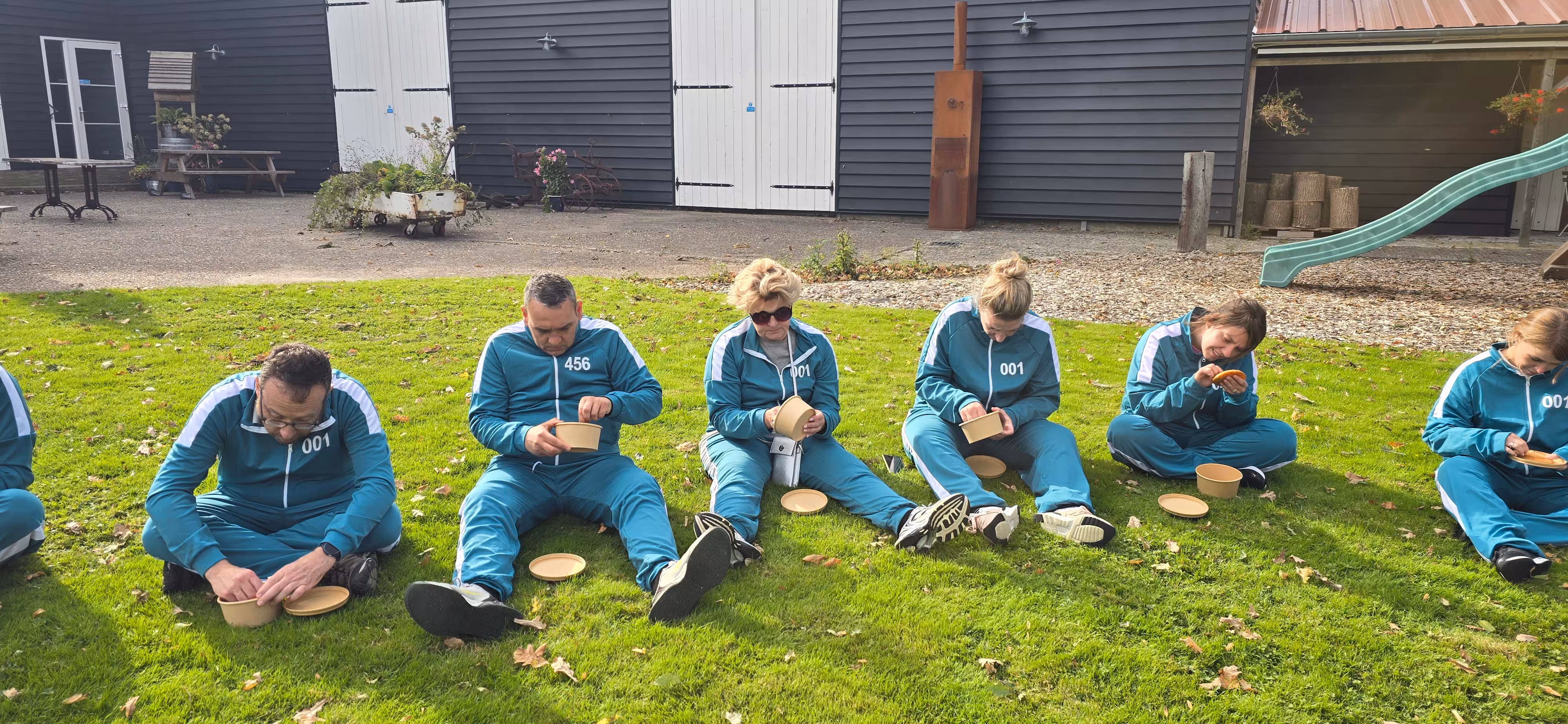 De deelnemers zitten op het grasveld in blauw outfit en proberen een koekje uit een bakje uit te snijden
