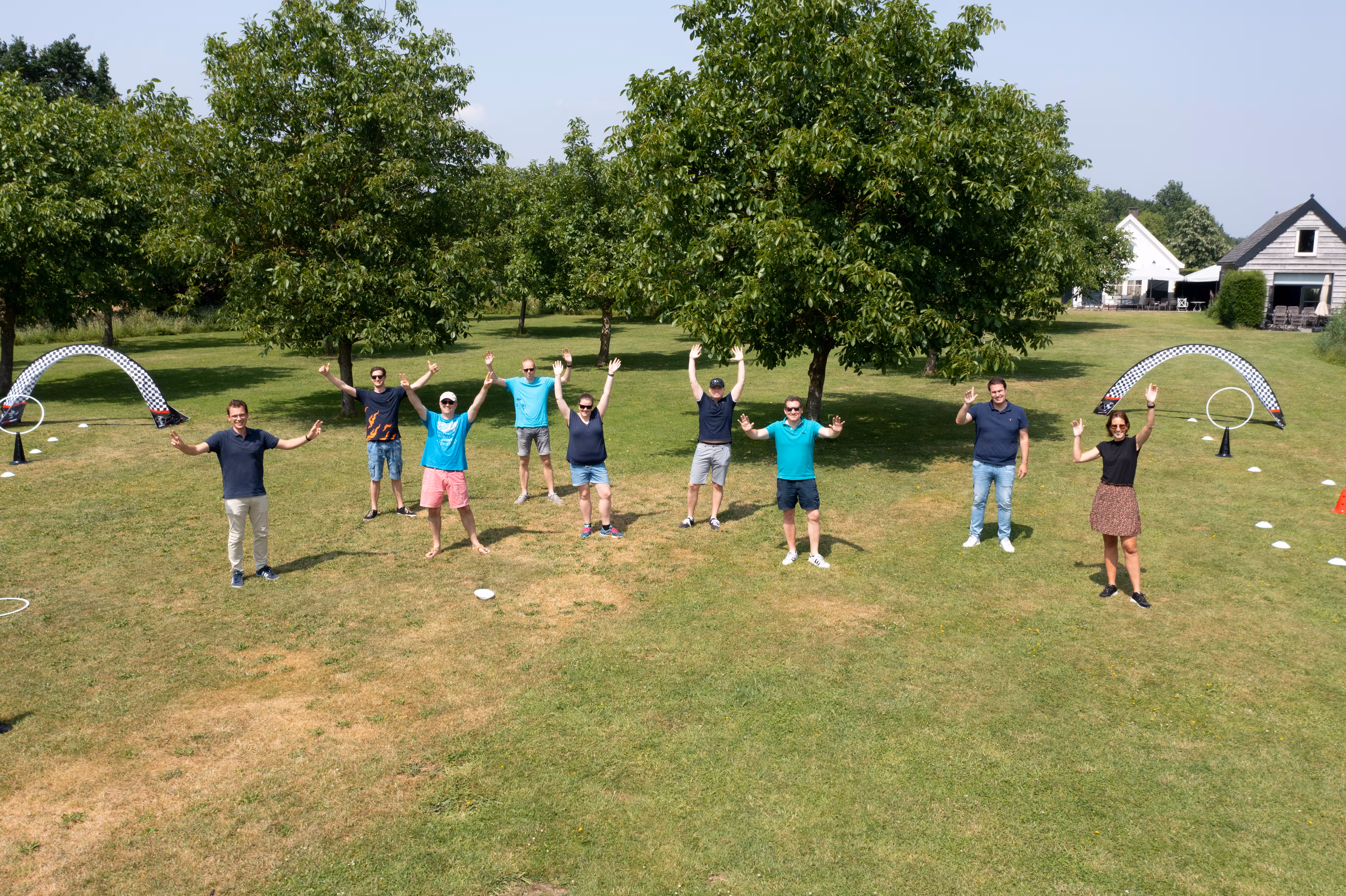 Groep deelnemers staat op een grasveld met armen omhoog tijdens een droneworkshop