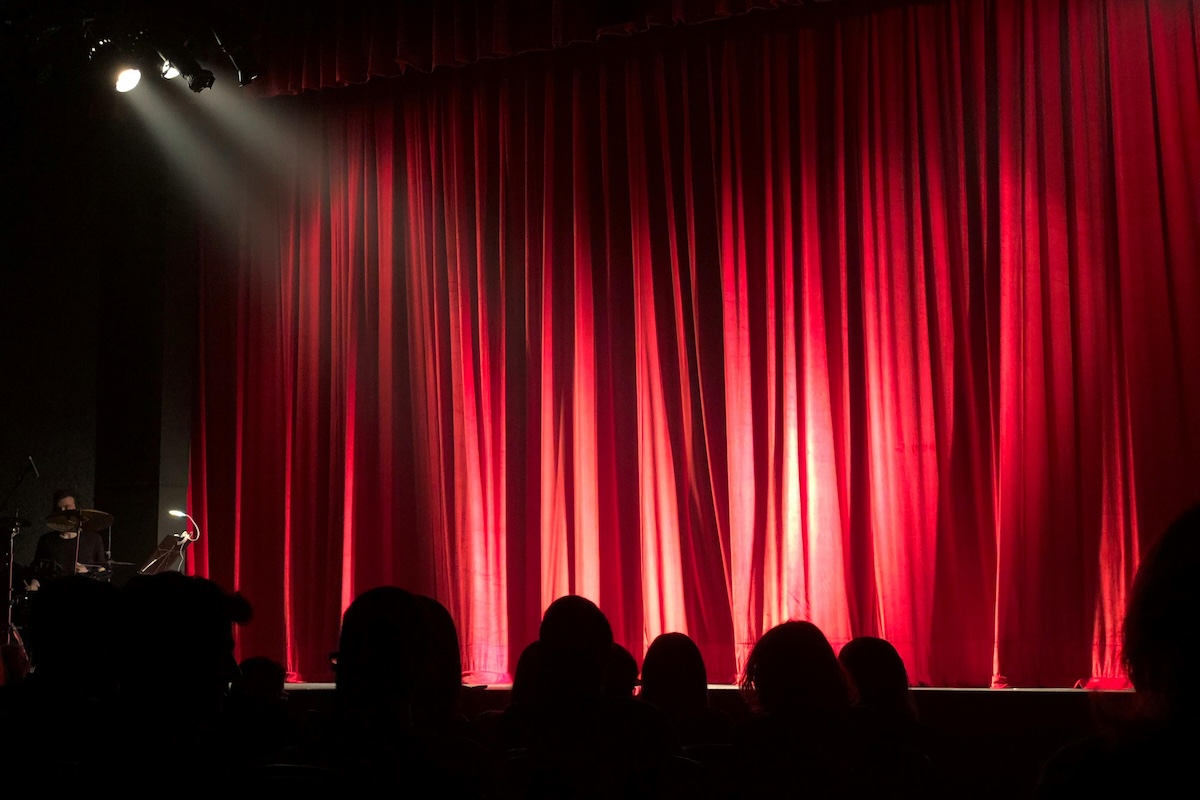 A red curtain hangs in a dark theater with a full audience.