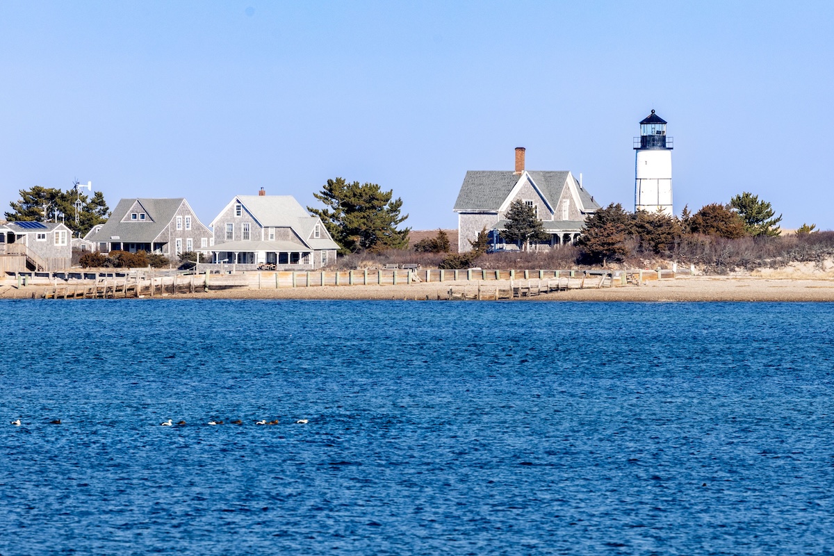 Picturesque Cape Cod lighthouse with traditional homes by the shoreline under a clear sky.