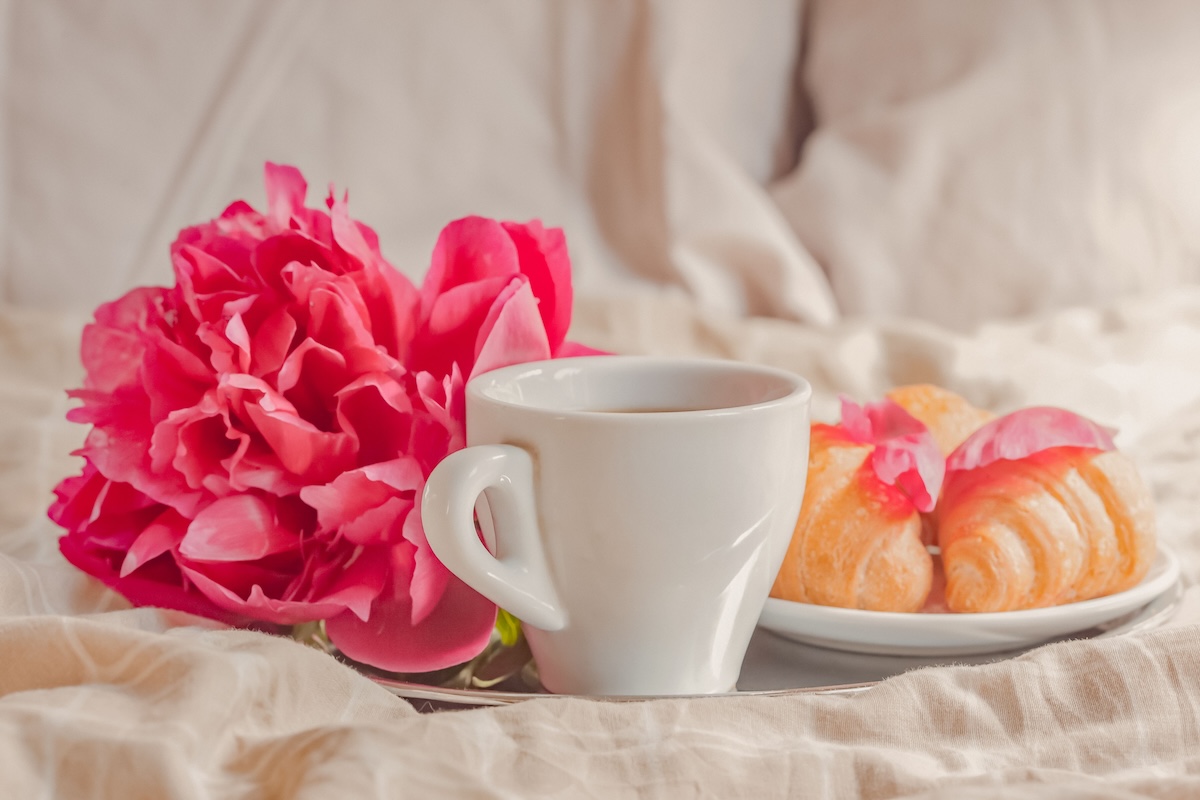 A warm and inviting setup of coffee, croissants, and a peony flower on a bed, perfect for a cozy morning.