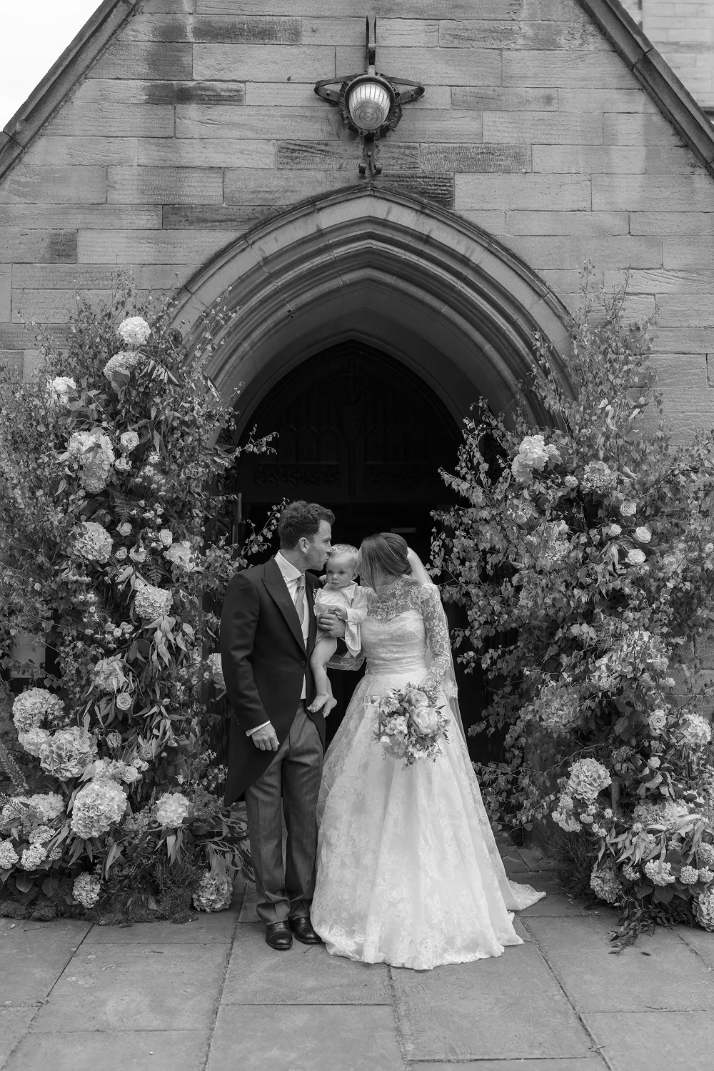 bride and groom outside church with their baby surrounded by flowers