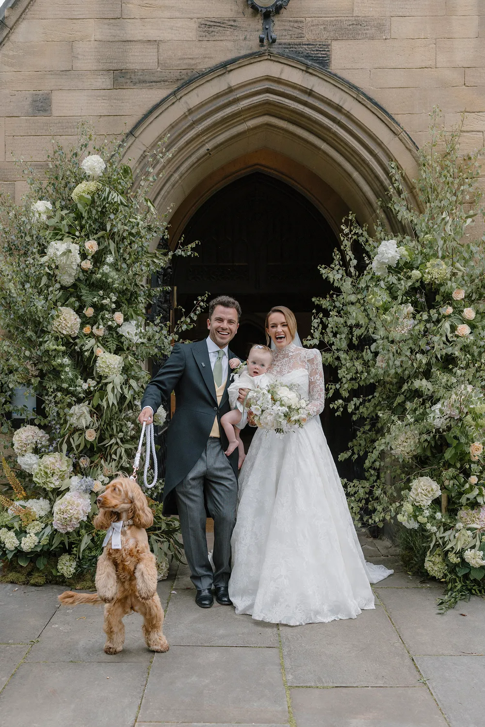 bride and groom with their baby and dog outside church surrounded by flowers
