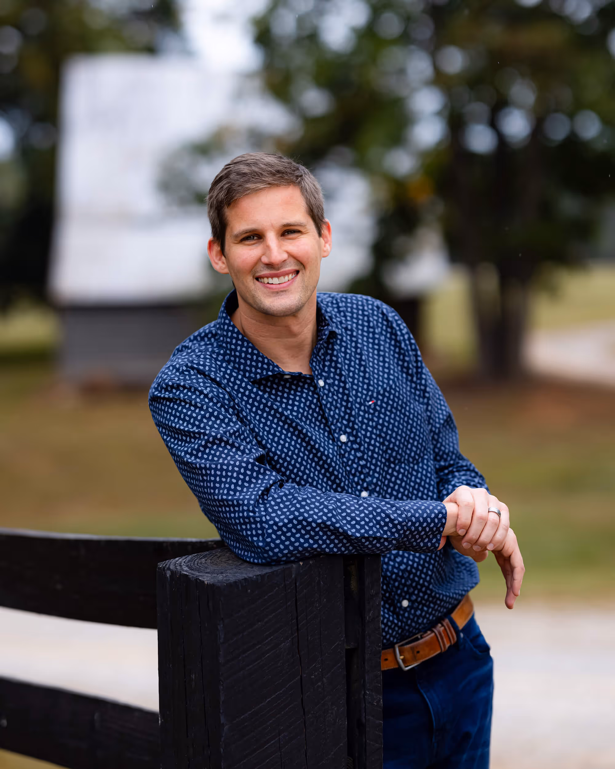 Smiling man in a blue patterned shirt leaning on a black wooden fence outdoors.