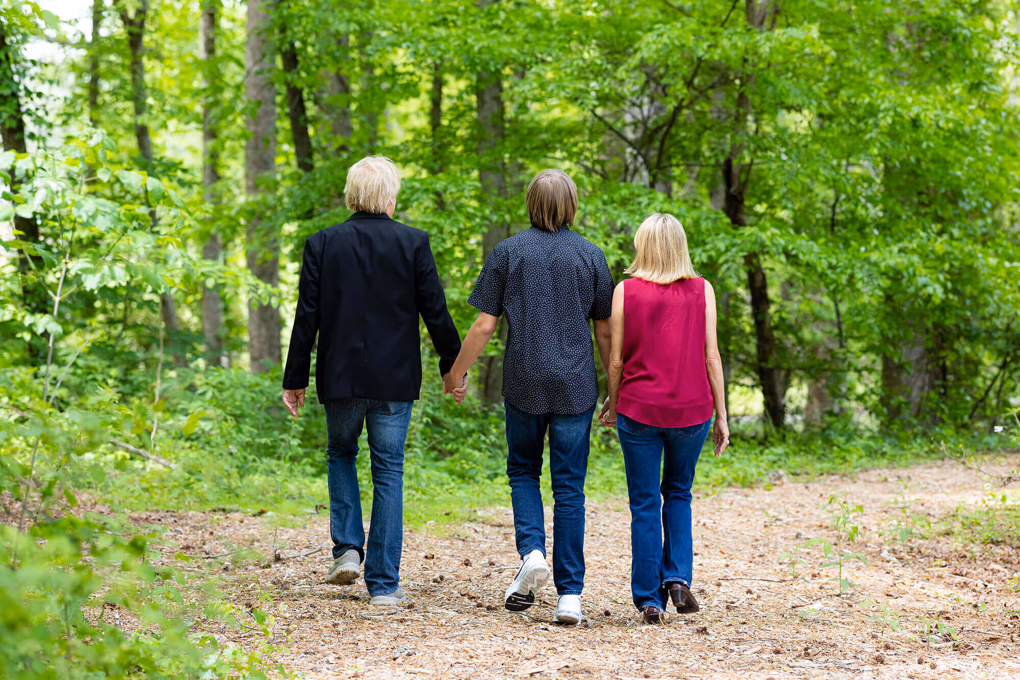 Three people walking hand in hand on a forest trail, surrounded by lush green trees.