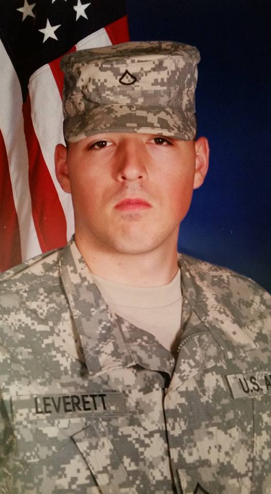 Portrait of a soldier in U.S. Army uniform with digital camo and hat, standing in front of the American flag.
