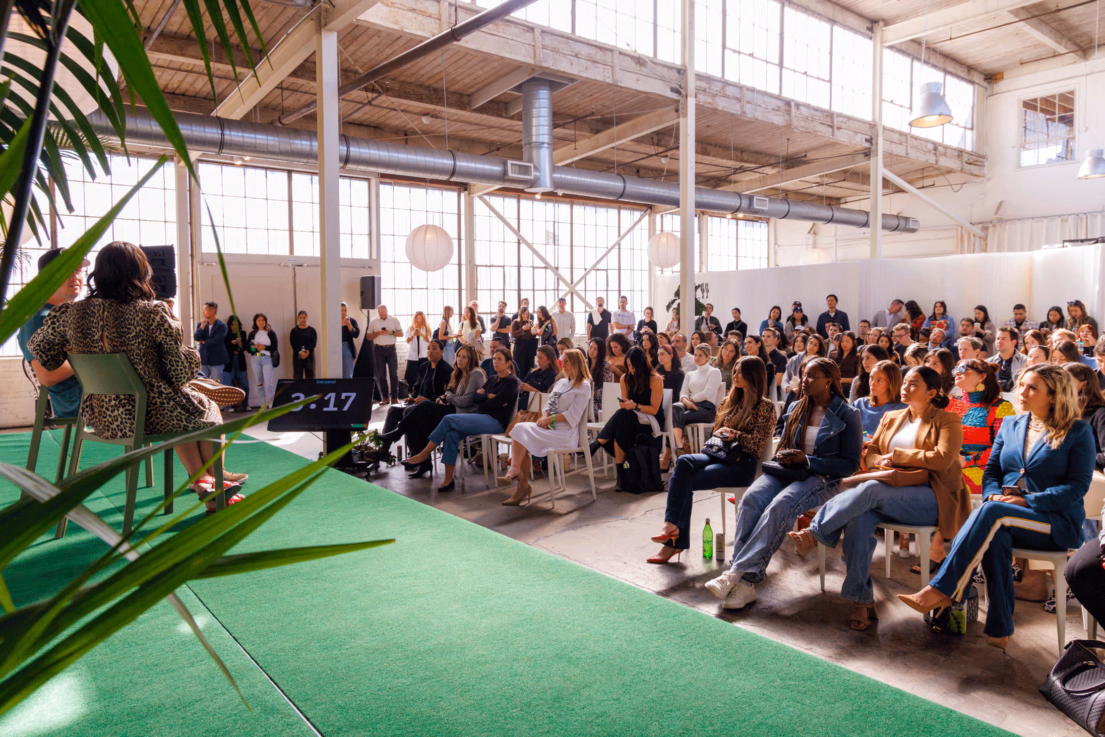 Audience seated indoors attentively listening to two speakers on a green-carpeted stage in a sunlit industrial-style room.