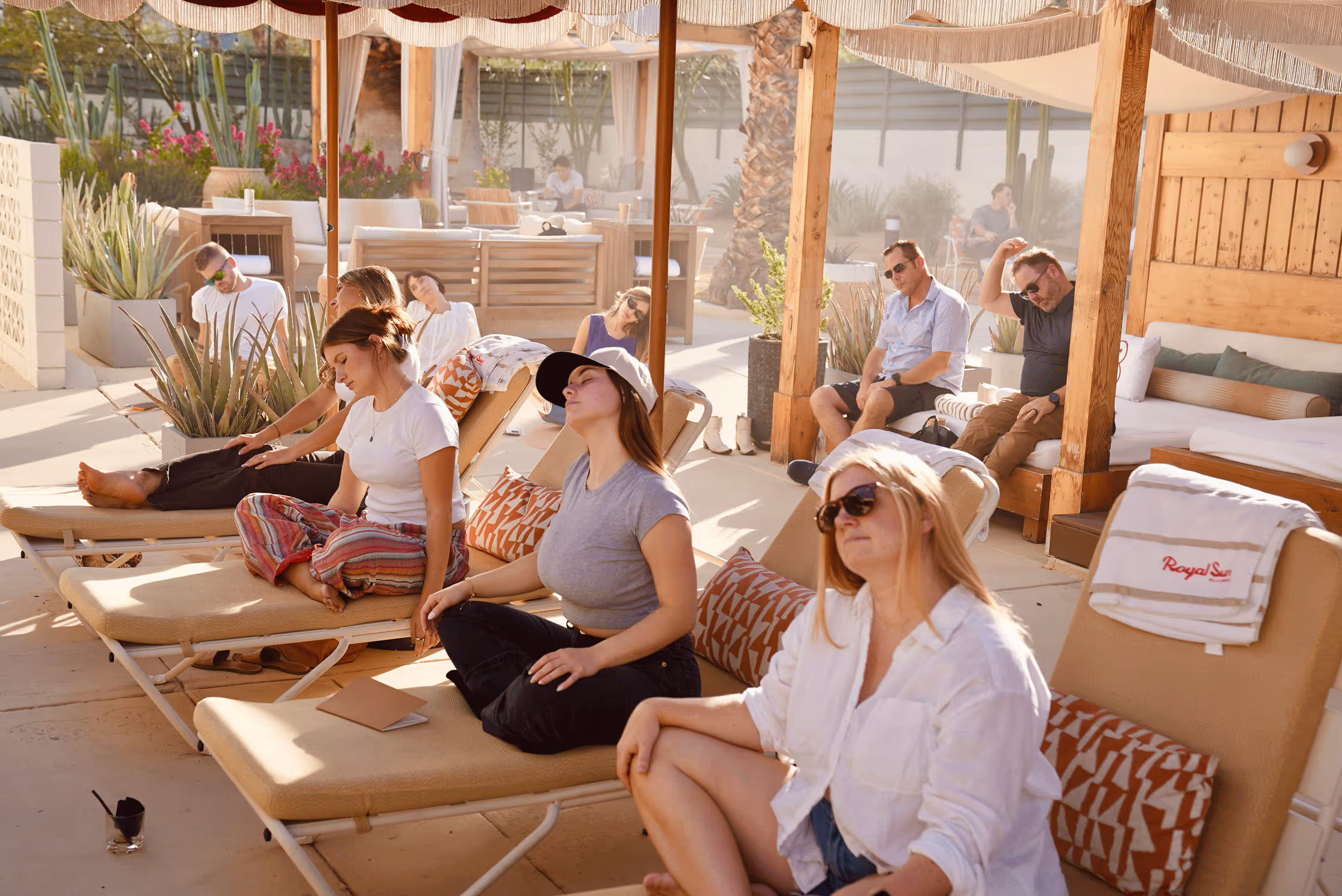 Group of people relaxing and meditating on lounge chairs under shaded cabanas by a pool during the daytime.