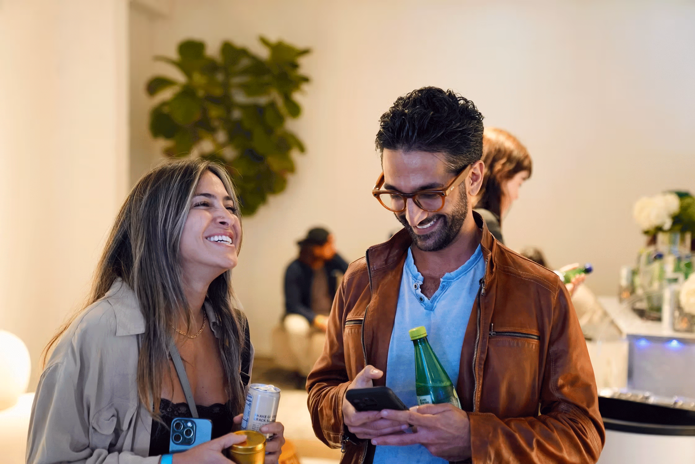 Two people smiling and holding drinks while looking at a phone at a social gathering indoors.
