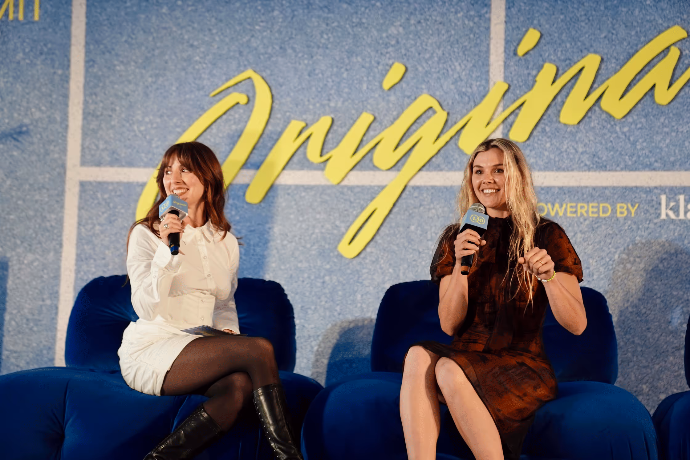 Two women sitting on blue chairs holding microphones, smiling during a panel discussion with the word 'Original' in large yellow script on a blue background behind them.