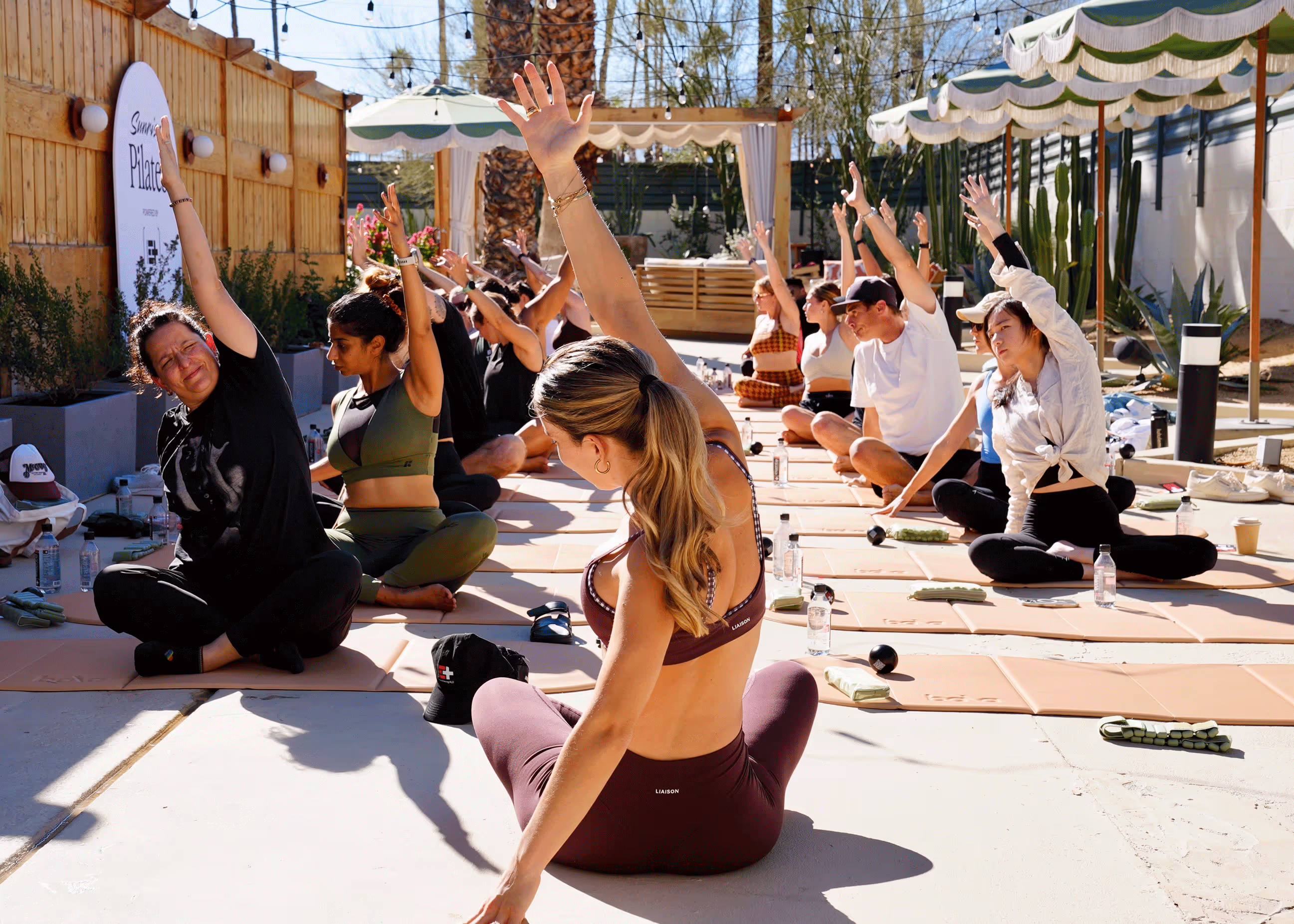 Group of people seated on yoga mats outdoors, stretching with one arm raised during a Pilates class under string lights and umbrellas.