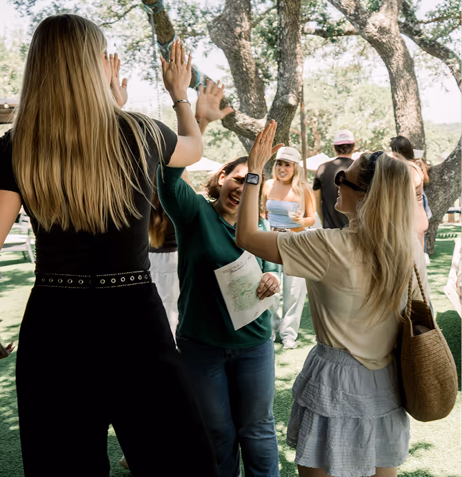 Three women outdoors exchanging high-fives with others socializing in the background under trees.