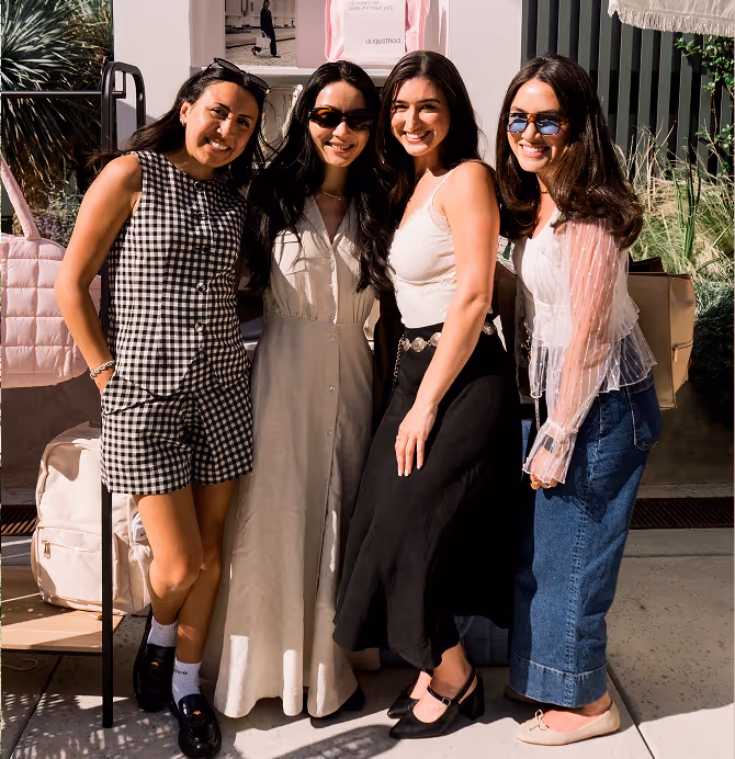 Four women standing close together outdoors, smiling at the camera, dressed in stylish casual outfits and sunglasses.