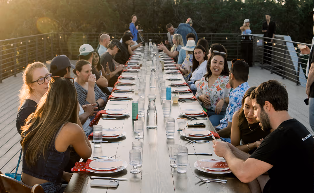 Large group of people seated at a long outdoor dining table during sunset, engaged in conversation.