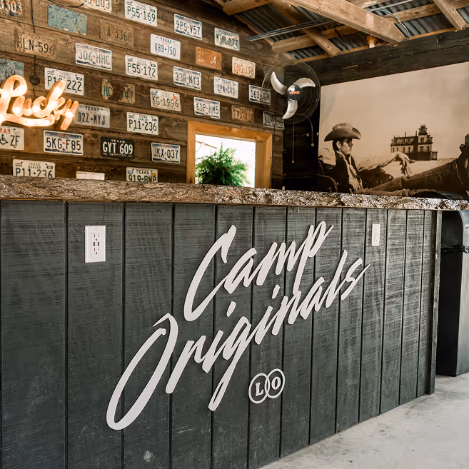 Interior of a rustic space with a black wooden counter displaying 'Camp Originals' in white letters, a wall decorated with vintage license plates, a neon sign reading 'Lucky,' and a black-and-white photo of a man in a cowboy hat.