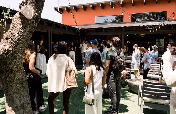 Group of people standing and listening outdoors near a modern building with string lights overhead.