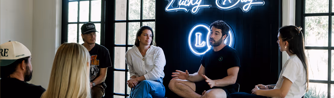 Five people engaged in a discussion in a room with large windows and a neon sign that says 'Lucky Day' on the wall.