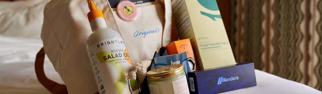 Cloth tote bag on a bed holding a bottle of Brightland salad oil, various skincare and wellness products, and a pink button badge.