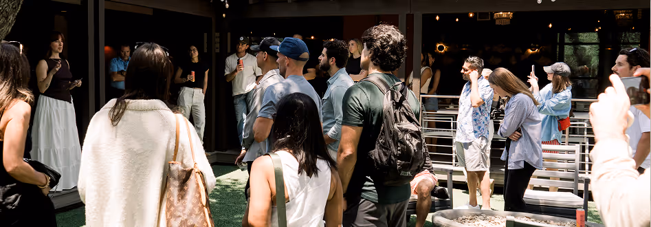 Group of people standing and listening attentively to a woman speaking in an outdoor setting with green artificial grass.