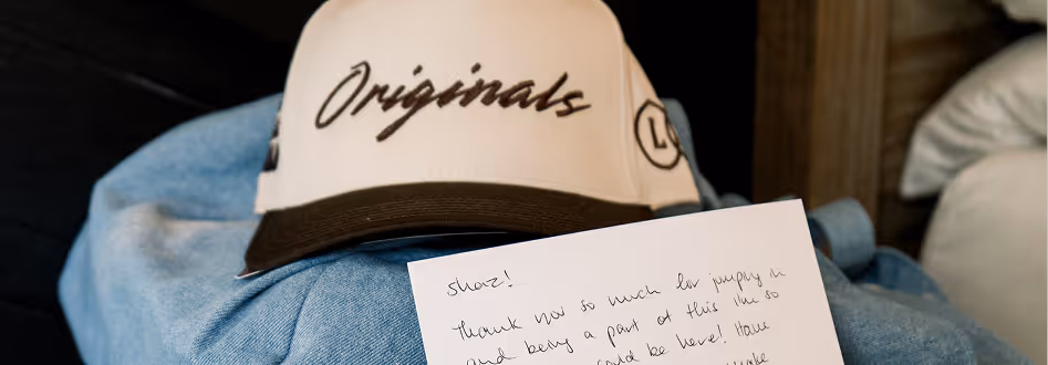 Handwritten note placed on blue fabric with a white and brown cap labeled 'Originals' in the background.