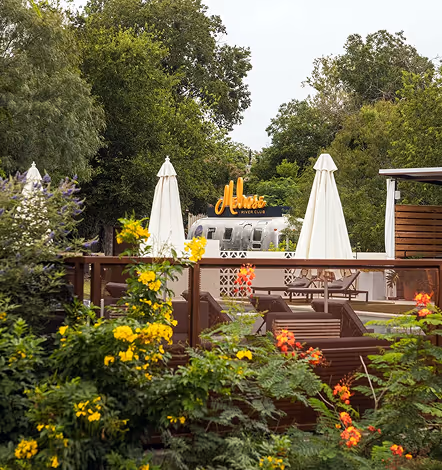Outdoor lounge area with white umbrellas, surrounded by greenery and colorful flowers, featuring an airstream trailer with a sign reading 'Moss River Club'.