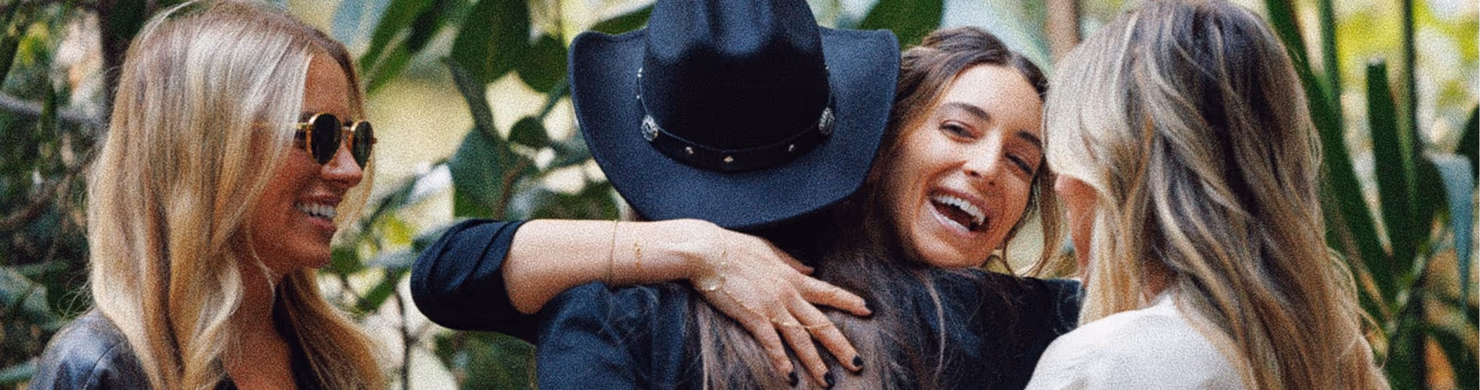 Three women smiling and embracing outdoors with greenery in the background.