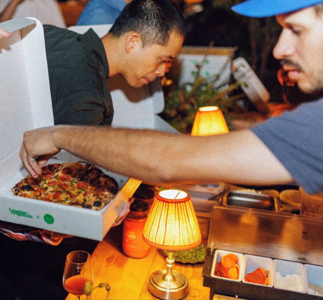 Two men sharing a pizza at a warmly lit table with a small lamp and condiment containers.