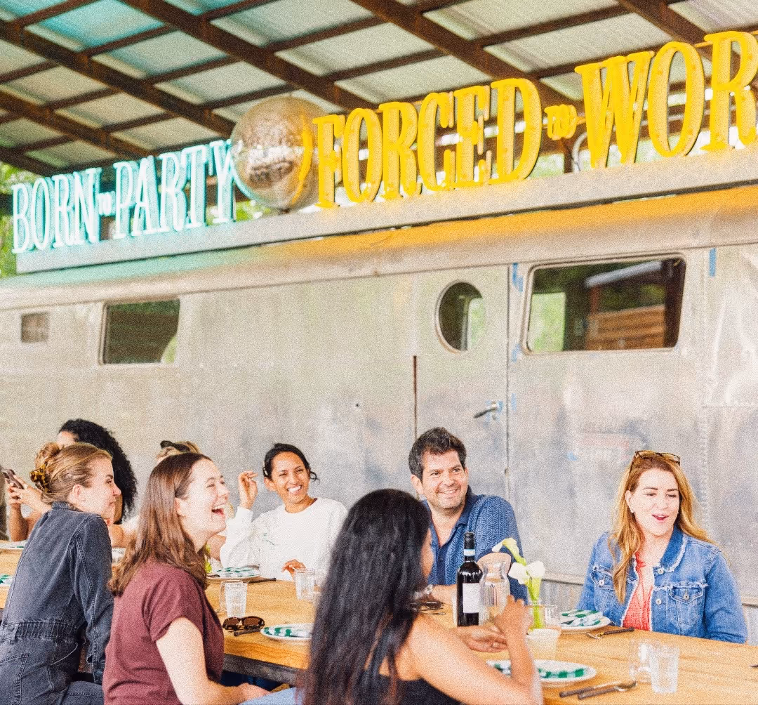 Group of people laughing and sitting at a wooden table with drinks and plates under a roof.