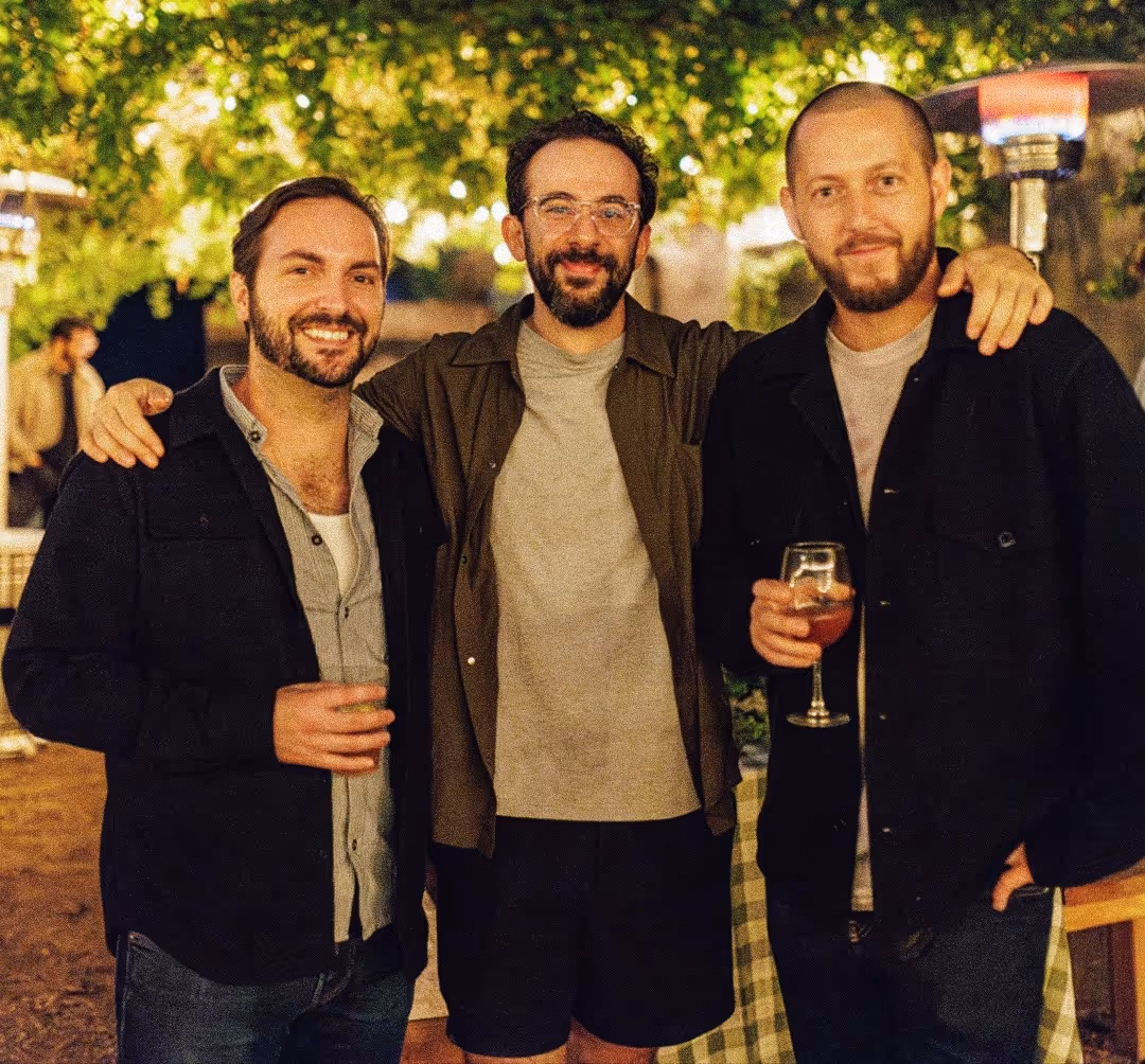 Three men smiling outdoors at night, two holding drinks, with trees and lights in the background.