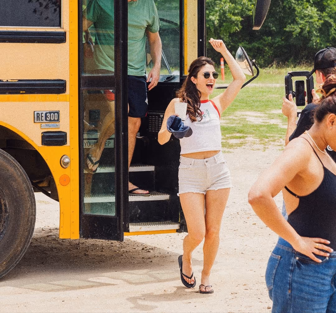 Woman in white shorts and tank top cheerfully exits yellow bus holding a drink and sunglasses.