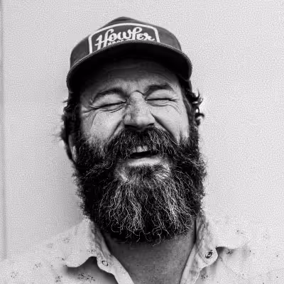 Bearded man with a hat smiling with eyes closed against a plain background.